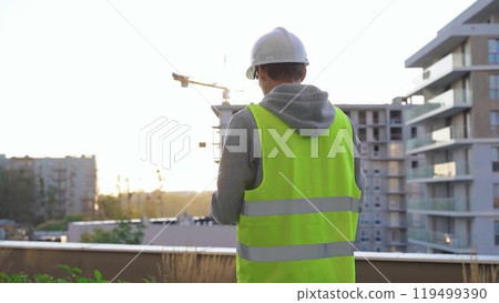 Man engineer with a white protective helmet and safety vest is writing on a clipboard while inspecting a construction site at sunset. Back view 119499390