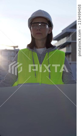 Female architect or engineer wearing safety hard hat and vest holding blueprint while inspecting a construction site at sunrise in early morning, front vertical view. Architecture concept Female architect or engineer wearing safety hard hat and vest holding blueprint while inspecting a construction site at sunrise in early morning, front vertical view. Architecture concept 119499408