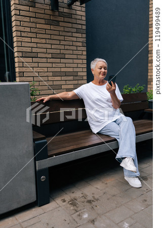 Vertical portrait of senior elderly 60s woman holding smartphone watching mobile video calling online, smiling looking at screen relaxing on city bench. Older grandmother using modern technology. 119499489
