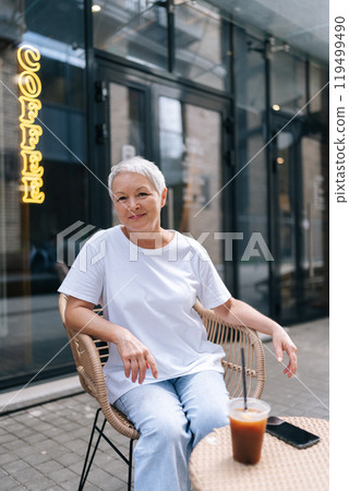 Charming elderly woman savoring iced coffee outside bustling cafe, appearing peaceful and at ease on sunny day, with smartphone close by to stay connected, smiling looking at camera. Charming elderly woman savoring iced coffee outside bustling cafe, appearing peaceful and at ease on sunny day, with smartphone close by to stay connected, smiling looking at camera. 119499490
