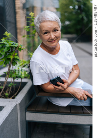 Vertical portrait of cheerful senior woman with short grey hair holding smartphone while sitting on urban bench outside, possibly enjoying moment of relaxation in city, smiling looking at camera. Vertical portrait of cheerful senior woman with short grey hair holding smartphone while sitting on urban bench outside, possibly enjoying moment of relaxation in city, smiling looking at camera. 119499497