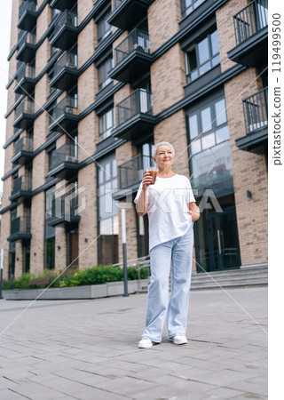 Vertical low-angle portrait of stylish senior woman drinking morning coffee outside modern city apartment, looking up with hope and contentment, enjoying break and savoring moment in retirement. Vertical low-angle portrait of stylish senior woman drinking morning coffee outside modern city apartment, looking up with hope and contentment, enjoying break and savoring moment in retirement. 119499500