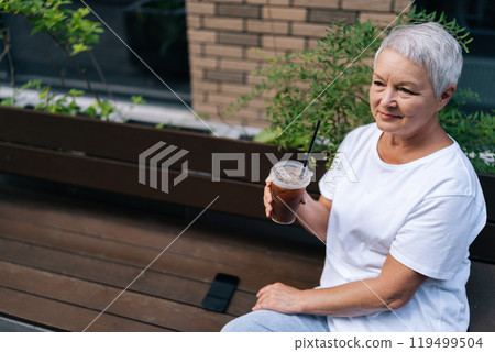 High-angle view of mature aged woman drinking iced coffee, looking content and relaxed in city. European gray-haired older female sitting on bench, smiling happily, enjoying free time in retirement 119499504