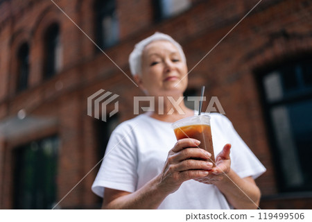 Low-angle view of gray-haired senior woman enjoying cold iced coffee on hot summer day in front of brick building, radiating happiness and relaxation, smiling looking at camera, selective focus. 119499506