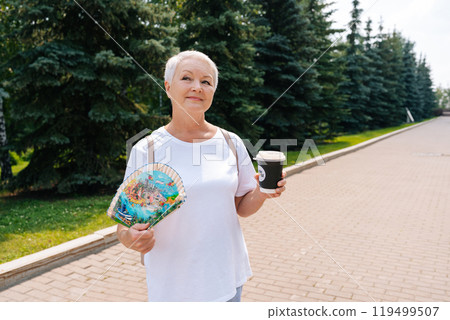 Portrait of elegant mature woman with short gray hair cooling down with fan while enjoying coffee on summer sunny day. European gray-haired older female walking in park, surrounded by green trees. 119499507