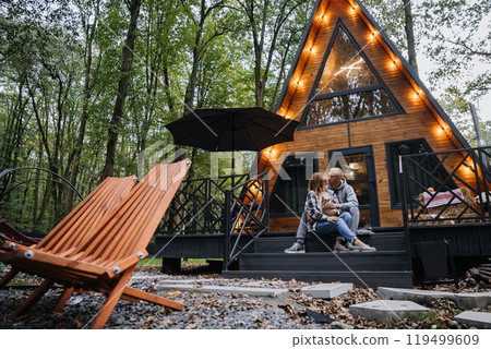 Cozy couple with cat enjoying peaceful moment at a wooden cabin in the forest, surrounded by nature Cozy couple with cat enjoying peaceful moment at a wooden cabin in the forest, surrounded by nature 119499609