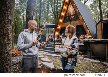 Couple preparing skewers for barbecue over open fire at cabin, enjoying autumn grilling together Couple preparing skewers for barbecue over open fire at cabin, enjoying autumn grilling together 119499611