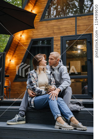 A loving couple sitting with their cat on wooden cabin steps, enjoying a cozy moment in nature 119499650