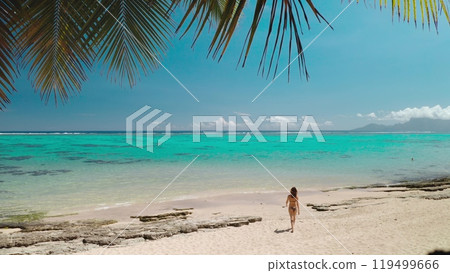 Enjoying the sunny weather on a pristine beach with turquoise water and palm trees, a young woman is walking on the white sand 119499666