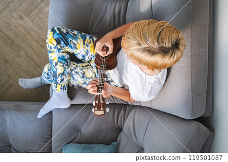Teen boy plays ukulele guitar on modern couch. Teen boy plays ukulele guitar on modern couch. 119501087