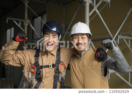 Two construction workers wearing safety harnesses and outdoor work are smiling and looking at the camera, making a fist pump 119502029