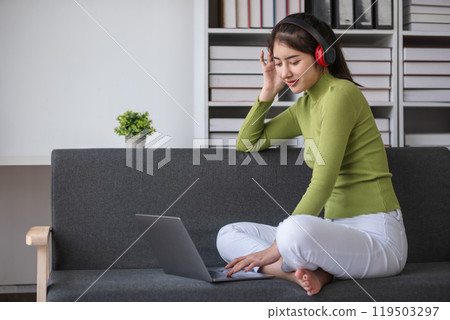 Young Woman Studying Online from Home on Laptop, Wearing Headphones, Sitting on Couch in Modern Living Room, Engaged in E-Learning, Remote Education, and Online Courses 119503297