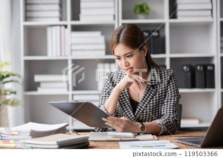 Young woman stressed at work, sitting at a desk with documents, holding a tablet, looking worried and overwhelmed in a modern office environment with shelves and plants in the background. 119503366