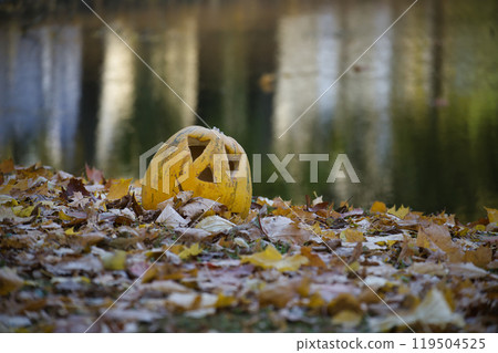 Carved pumpkin sits among vibrant fallen leaves near a tranquil pond 119504525