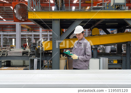 Young asian worker check tasks while wearing a safety helmet and gloves, surrounded by machine and material. Work atmosphere in metal sheet manufactory. Young asian worker check tasks while wearing a safety helmet and gloves, surrounded by machine and material. Work atmosphere in metal sheet manufactory. 119505120