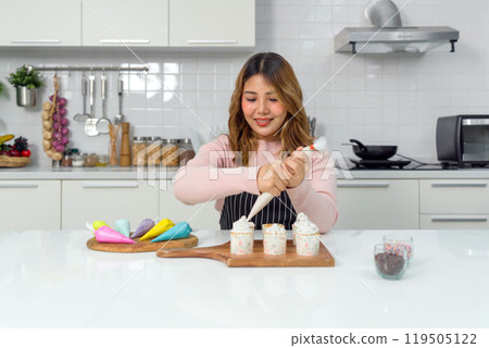 Young asian woman in an apron using a piping bag, pipe the icing onto the cupcake, decorating cupcake on a wooden board, adding a colorful touch to the cupcakes' presentation. Young asian woman in an apron using a piping bag, pipe the icing onto the cupcake, decorating cupcake on a wooden board, adding a colorful touch to the cupcakes' presentation. 119505122