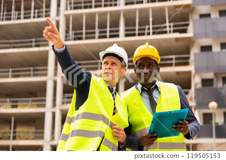 Young civil engineer discussing a construction plan with a colleague shows him something 119505153