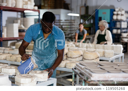 Slip casting - african american worker pours slip mass from jug into plaster mold in pottery factory Slip casting - african american worker pours slip mass from jug into plaster mold in pottery factory 119505268