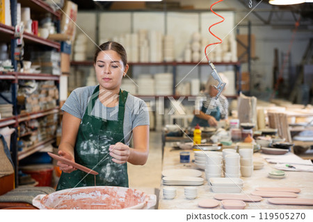 Young female worker in pottery factory lowers fired clay plates and cups into container with glaze Young female worker in pottery factory lowers fired clay plates and cups into container with glaze 119505270
