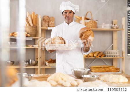 Portrait of baker holding tasty hot bread and baguettes in hands in kitchen Portrait of baker holding tasty hot bread and baguettes in hands in kitchen 119505512
