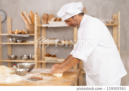 Male baker stands at work bench, kneading and shaping dough to make bread, croissants and baguettes 119505561