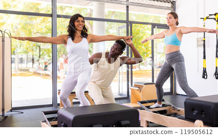 Attentive African American male instructor helping two positive women performing set of pilates Attentive African American male instructor helping two positive women performing set of pilates 119505612