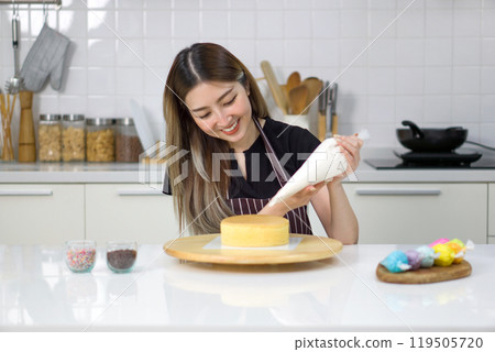 Young woman in an apron using a piping bag, pipe the icing onto the cake, creating a decorative pattern on the top of a round, light-colored cake that's placed on a wooden turntable or cake stand. Young woman in an apron using a piping bag, pipe the icing onto the cake, creating a decorative pattern on the top of a round, light-colored cake that's placed on a wooden turntable or cake stand. 119505720