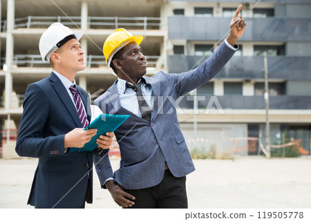 Two architects inspect the construction of new building. One of them points his hand to side Two architects inspect the construction of new building. One of them points his hand to side 119505778