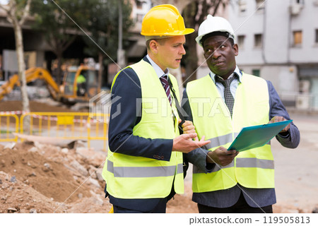 Portrait of two engineers in helmet and yellow vests. Indicate finger to side 119505813