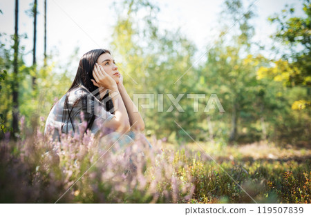 A young woman sits peacefully in a sunlit forest clearing, enjoying nature  119507839
