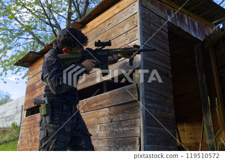 a young sniper aims at a house from the woods 119510572