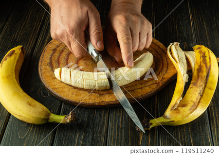 A cook cuts a ripe banana with a knife on a wooden cutting board to prepare a fruit dessert for breakfast 119511240