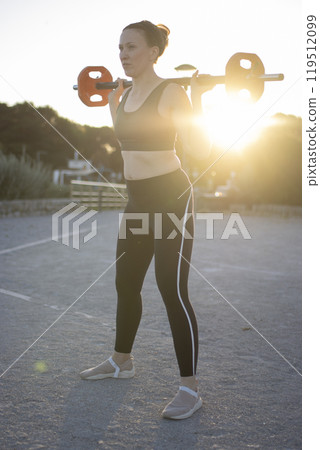 Young woman doing squats outdoors with weights at sunset 119512099