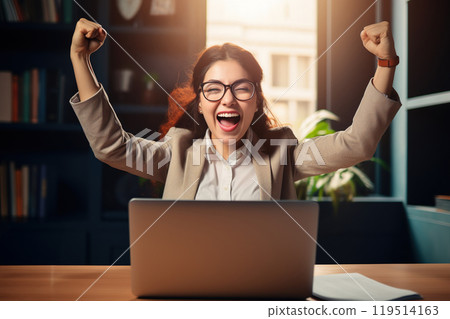 Happy businesswoman in her office, sitting at her desk with a laptop and shouting YES with hands up 119514163