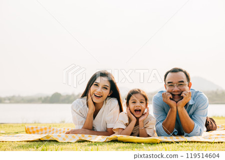 Asian family father mother and daughter lying on blanket spring meadow field grass in park cheerful sweet kid girl, parents and child picnic enjoying outdoors sunset time together, Family relaxation 119514604