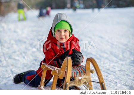Happy Little boy kid portrait wear overall enjoy having fun sledding vintage wooden sledge on snowy hill in city park outdoors at winter holiday. Smiling child riding sport bob outside 119516406