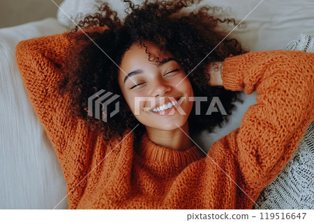 A young woman in a cozy orange sweater relaxes with a joyful smile on a comfortable couch in a A young woman in a cozy orange sweater relaxes with a joyful smile on a comfortable couch in a 119516647