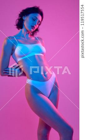 Woman stands in white lingerie, elegantly posing with her body in vibrant neon hues of blue and pink, against gradient background. 119517554