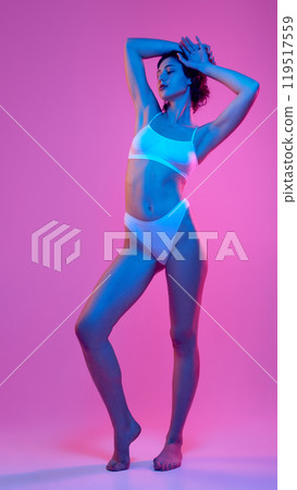 Woman posing elegantly with her arms raised, gracefully accentuating her curves in neon light against vibrant, colorful backdrop. Woman posing elegantly with her arms raised, gracefully accentuating her curves in neon light against vibrant, colorful backdrop. 119517559