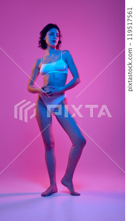 Full-body portrait of woman in white underwear, standing gracefully against pink and blue-lit backdrop, highlighting her toned body and confident pose. 119517561