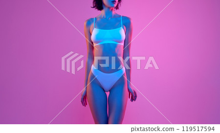 Fit woman standing with hands at her sides, wearing white underwear and posed against pink background, lit in neon hues of blue and pink. Fit woman standing with hands at her sides, wearing white underwear and posed against pink background, lit in neon hues of blue and pink. 119517594