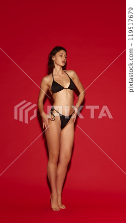 Toned young woman in bikini, confidently posed with subtle elegance against vivid red background. Swimwear advertising. Toned young woman in bikini, confidently posed with subtle elegance against vivid red background. Swimwear advertising. 119517679
