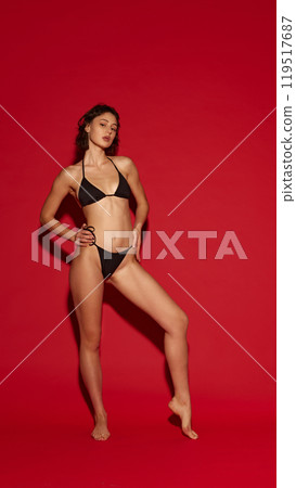 Young attractive woman poses gracefully in black bikini, showing her athletic body forms against vivid red background. Young attractive woman poses gracefully in black bikini, showing her athletic body forms against vivid red background. 119517687