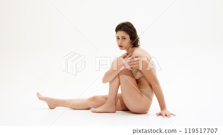 Portrait of young woman sitting on floor in neutral lingerie, embodies balance between health, beauty against white studio background. 119517707