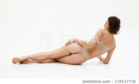 Rear view of young woman in underwear lying against white studio background, Her toned figure and soft glow, model showcases results of self-care Rear view of young woman in underwear lying against white studio background, Her toned figure and soft glow, model showcases results of self-care 119517716