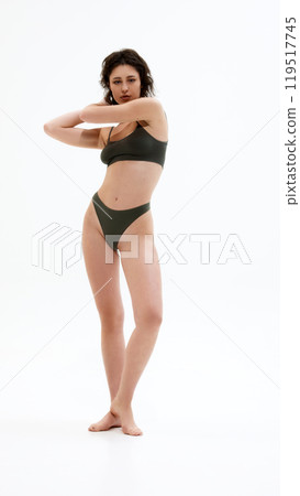 Full-length photo of young attractive woman posing in dark green lingerie and looking at camera, showing confidence against white studio background. 119517745