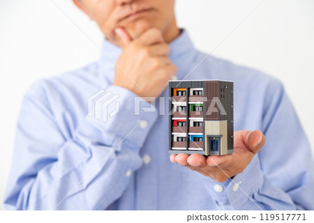 A middle-aged man holding a model of an apartment building and worrying 119517771