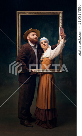 Couple in vintage-inspired clothing posing with golden frame while taking modern-day selfie against dark vintage background. Couple in vintage-inspired clothing posing with golden frame while taking modern-day selfie against dark vintage background. 119517896