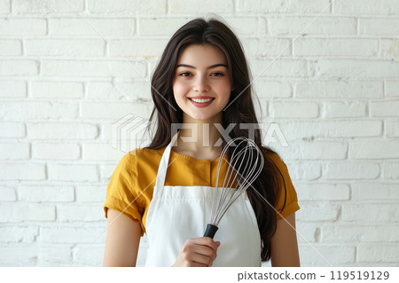 Young woman in an apron holding a whisk, smiling cheerfully in a bright kitchen with white brick 119519129