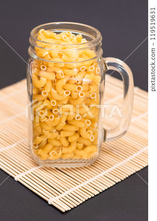 Uncooked Chifferi Rigati Pasta in Glass Jar on Bamboo Mat on Black Background 119519631